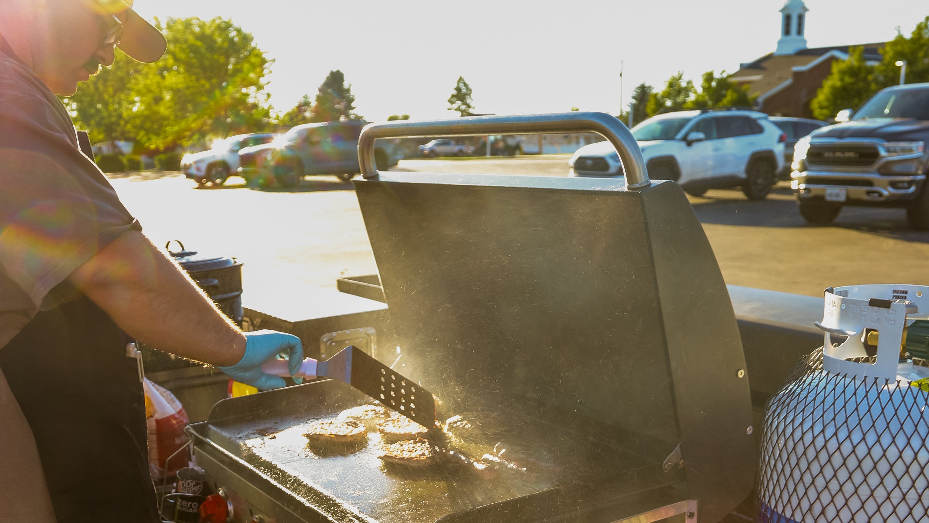 Person grilling outdoors with a propane tank and cooler nearby.